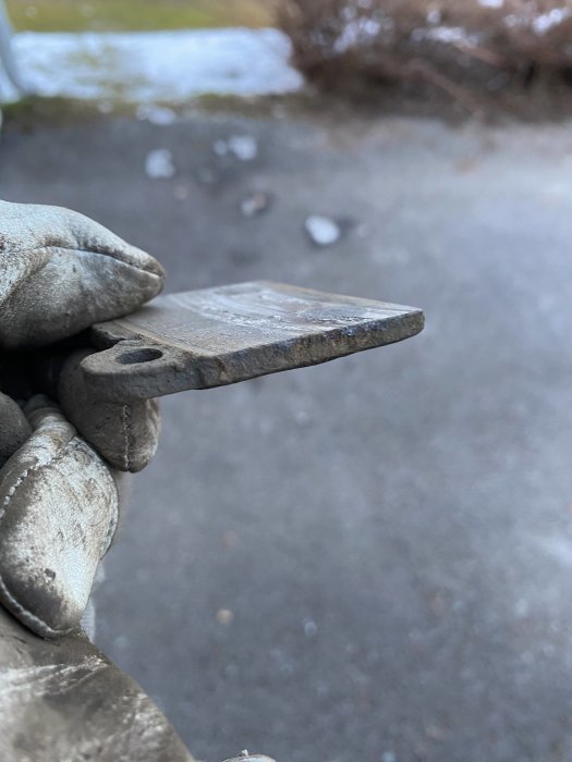 Hand i arbetshandske håller stålbult eller spik nära kamera; suddig utomhus bakgrund.