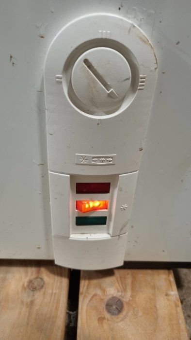 Smutsig termostat med upplysta röda och gröna lampor, kontrollhjul upptill, placerad på vägg.