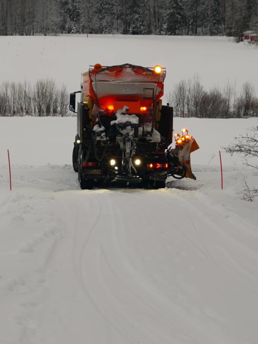 Snöig väg med en snöplogsbil i arbete, upplyst, röda avspärrningspinnar synliga, träd i bakgrunden.