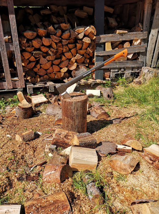 Yxa fast i trädstubbe framför vedstapel, kluvna vedträn och spån på marken.