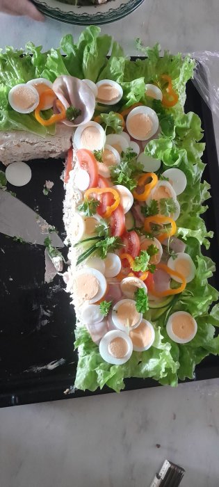 En smörgåstårta med ägg, skinka, sallad, tomat, gurka och persilja på ett svart fat.