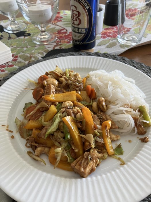 En tallrik med asiatisk mat, grönsaker, cashewnötter och risnudlar, intill en ölburk.