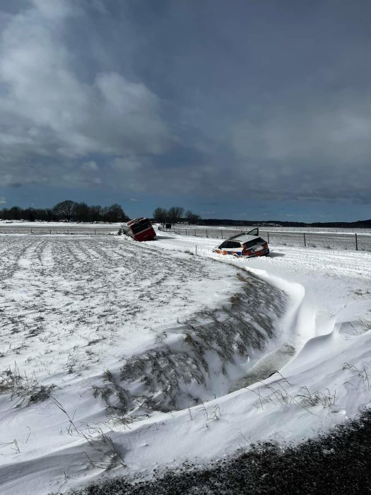 Två fordon i diket bredvid snöig väg, molnig himmel, vinterlandskap, vindskapad snödriva.