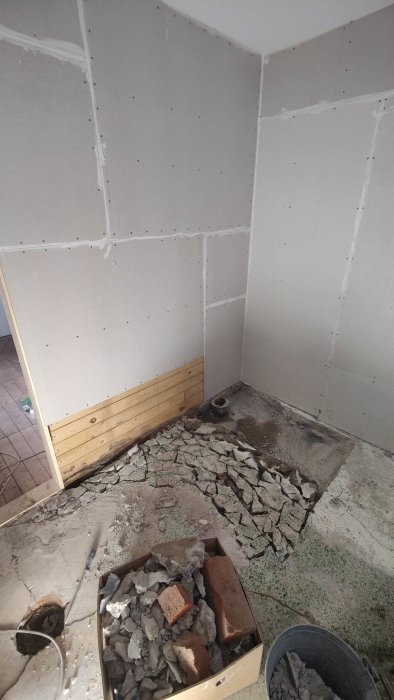 Hörn av ett rum under renovering, gipsväggar uppsatta, trasigt golv med skräp och tegelstenar.