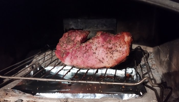 Ett stort köttstycke grillas, kryddat, över öppen eld i rökig miljö.