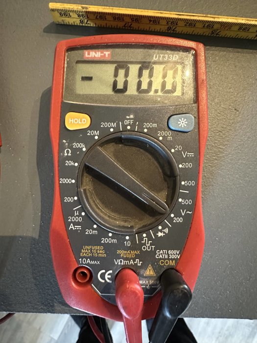 Digital multimeter, UNI-T märke, mäter -0.01 volt, testledningar anslutna, bakgrund av verktygsbänk.