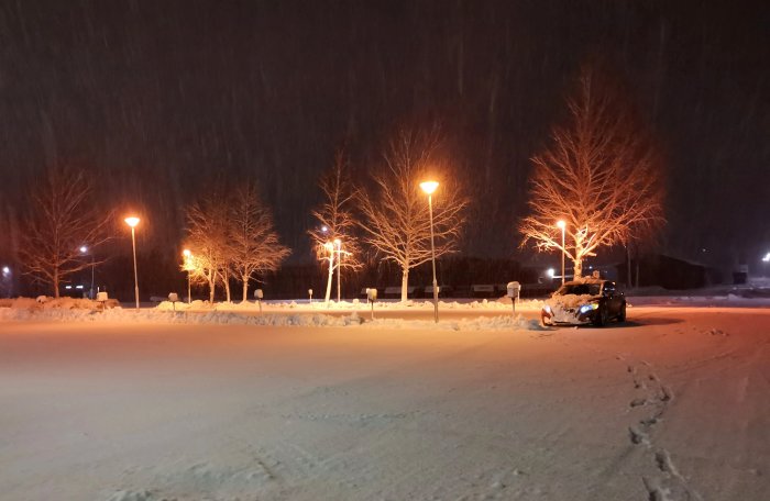 Ett parkerat fordon omgivet av snö, gatlyktor lyser upp, fotspår i snön, natt, snö faller.