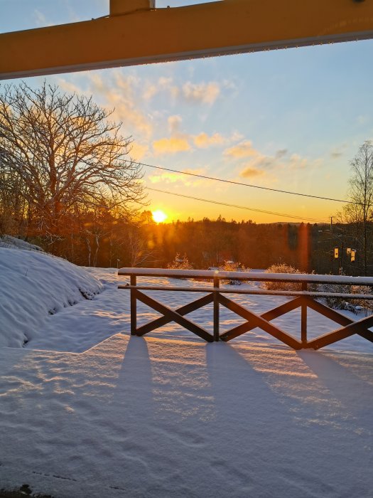 Vinterlandskap vid solnedgång, snö, trädstammar, moln, himmel, skuggor, räcke, och en balkong.