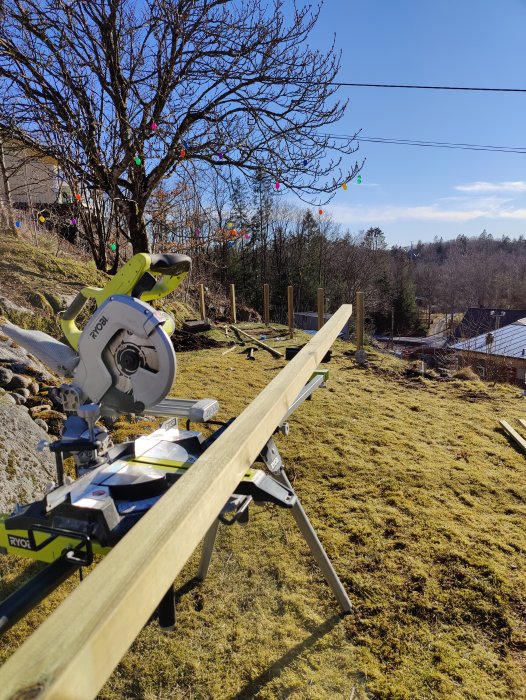 Elektrisk såg på arbetsbänk i solig trädgård med färgglada ljusslingor och barträd. Byggprojekt eller renovering pågår.