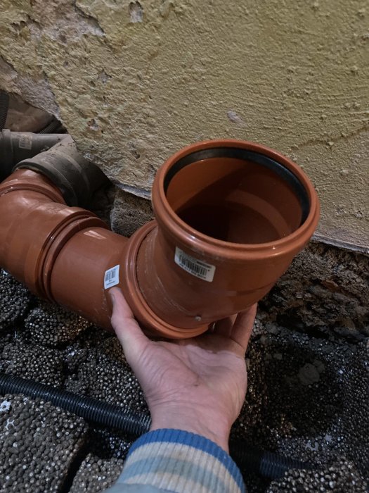 Hand håller ett terrakotta avloppsrör, gammalt murat vägg och grusig mark, arbetsmiljö.