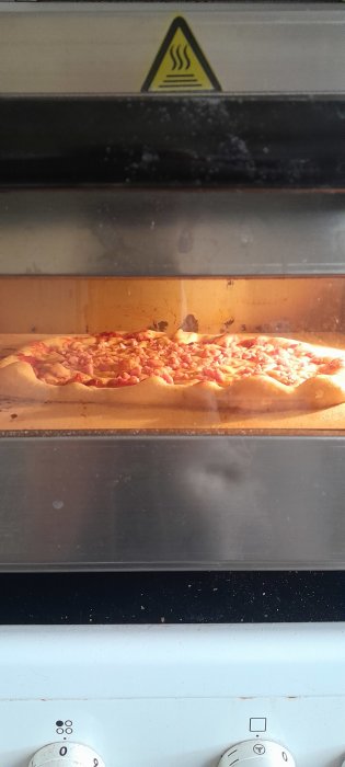 Pizza tillagas i ugn, synliga toppings, bakplåtspapper under, varningsskylt för värme ovanför.