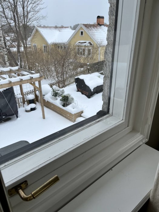 Vy genom ett fönster mot en snötäckt trädgård och gult hus.