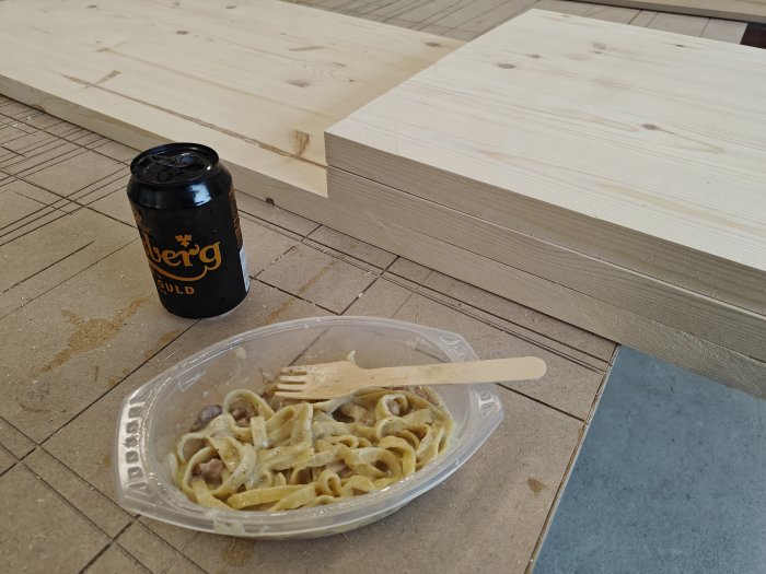En burk dryck, halväten pasta i plastbehållare med trägaffel på byggarbetsplats, träplankor i bakgrunden.