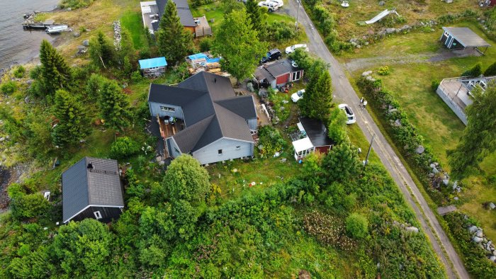 Flygbild över bostadshus vid vatten, grönska, fordon, båt vid brygga, väg och byggnader.