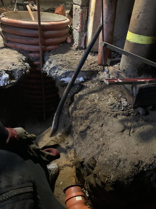 Grävplats inomhus, synliga rör och verktyg, kaotisk miljö, underjordiskt arbete, pågående renovering eller reparation.