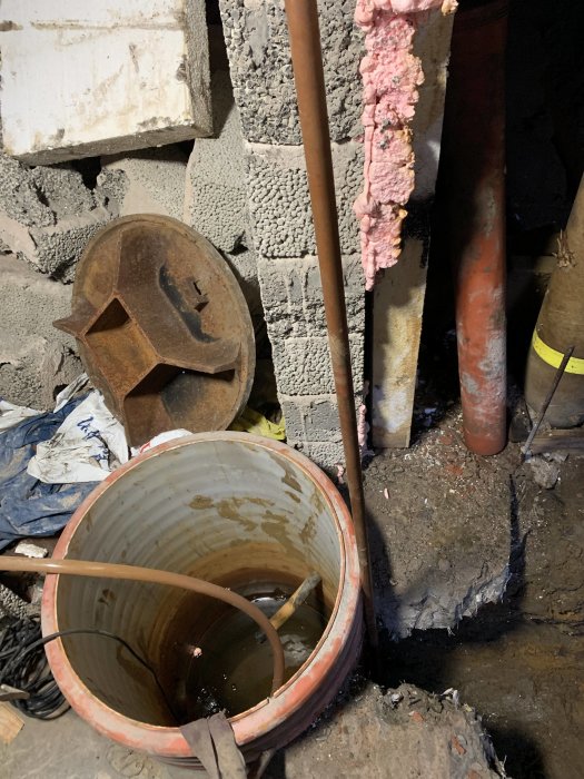 Rörigt källarutrymme med byggmaterial, isoleringsskum, hjälm och tom hink.