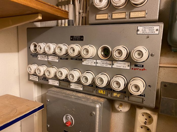 Äldre säkringsskåp med porslinsäkringar, några etiketter, en strömbrytare och vägguttag.