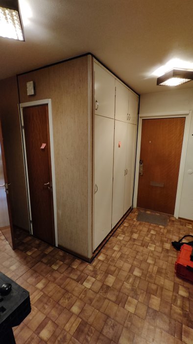 Ett inomhusutrymme med skåp, dörrar och brunmönstrat golv. Anteckningar på skåpdörrarna, väska på golvet.