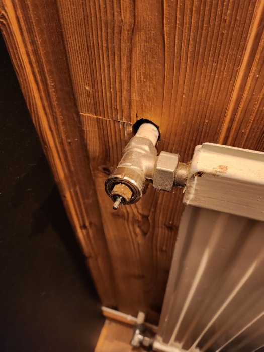 Radiatorventil som sitter fast i en träpanelvägg med synlig skada runt hålet.