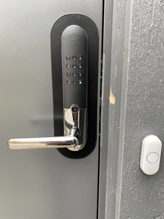 Svart dörr med elektroniskt kodlås, dörrhandtag och en Honeywell Home-dörrklocka.