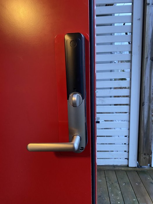 Röd dörr med digitalt lås och handtag, träaltan och vita persienner syns i bakgrunden.