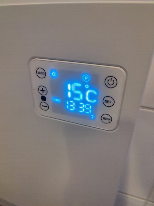 Digital termostat visar 15 grader Celsius med tid 13:34, placerad på vit vägg.