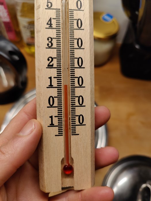 Trätermometer hållen i hand visar cirka 21 grader Celsius med köksredskap i bakgrunden.