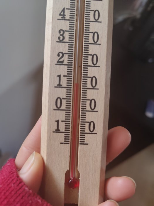 Trähållare med termometer visar cirka 20 grader Celsius. Hand håller termometer. Inomhusbelysning. Närbild.