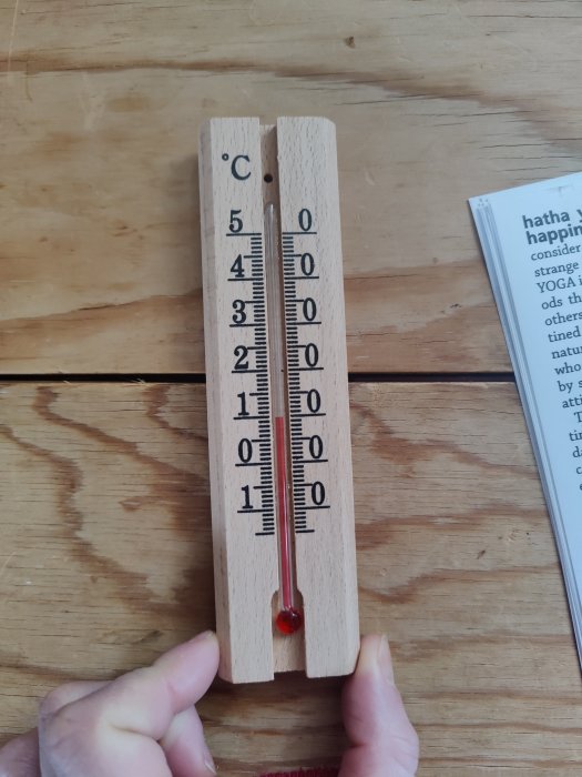 Termometer i trä visar temperatur i Celsius på träbord; hand håller i den.