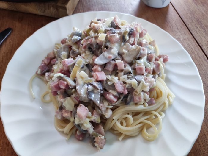 Pasta med krämig sås, skinka, champinjoner, serverad på vit tallrik, matlagning, måltid, hemlagat, köksbord.