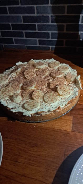 En banoffeepaj med vispgrädde, bananskivor och chokladströssel på ett mörkt träbord mot tegelväggsbakgrund.