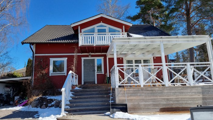 Rött tvåvåningshus med balkong, snö, carport, trädgårdsmöbler och blå himmel.