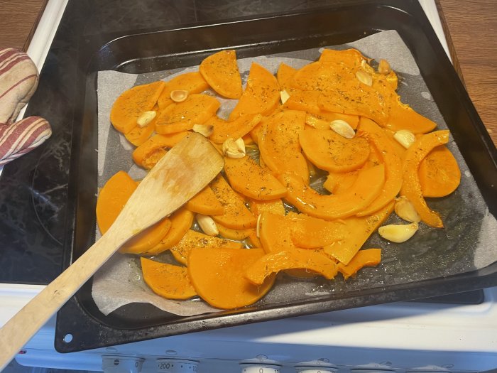 Skivad butternutpumpa kryddad med vitlök på bakplåt, redo för ugnsrostning. Träspatel och grytlapp syns.