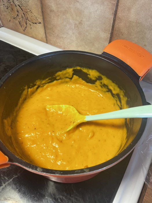 En kastrull med tjock, orange pumpasoppa och en vit slickepott i ett kök.