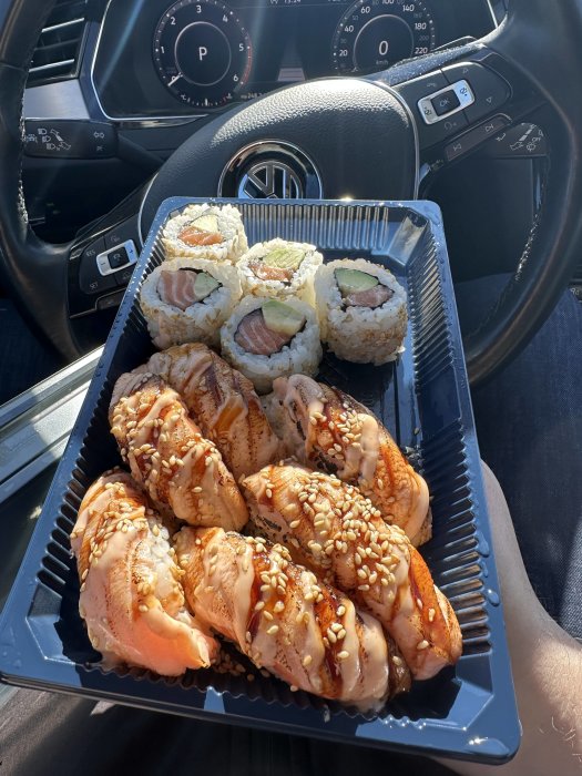 Sushi i bil, rullar med lax, sesamfrön, avokado, på någons knä, ratten synlig.