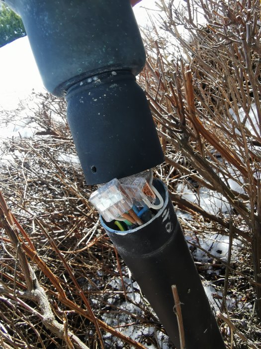 Öppen lampstolpe visar elanslutningar, omgiven av snötäckta buskar under dagsljus.