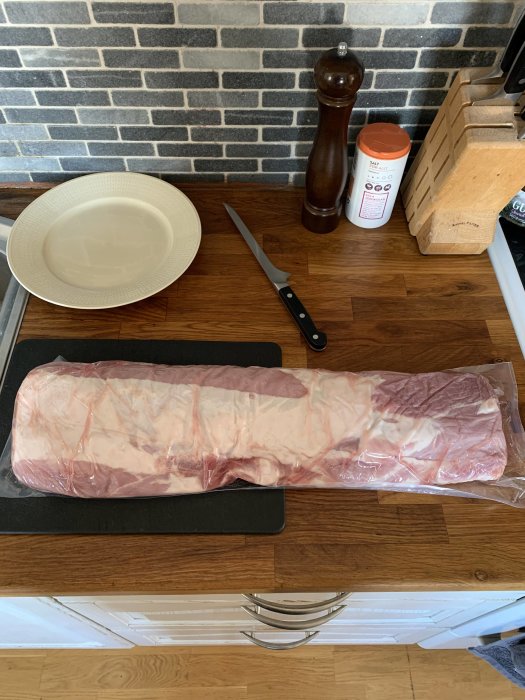 Ett paket rått kött på skärbräda, omgiven av kniv, kryddkvarn, tallrik och salt på köksbänk.