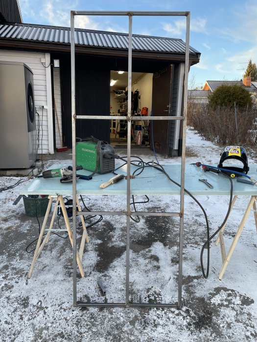 Metallram framför verkstad, verktyg och material på bord, snöfläckar på marken, öppen dörr i bakgrunden.