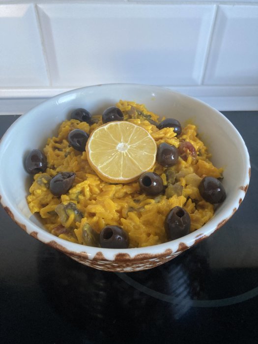 En skål med gyllengul risrätt, svarta oliver och citronskiva, vit kakelbakgrund, upplagd på mörk yta.