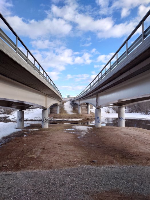 Två parallella broar över en isig flod, klar himmel, snöspår och lite graffiti på pelarna.