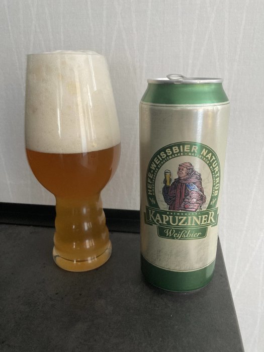 Ölglas fyllt med skummande öl bredvid en ölburk med etikett "Kapuziner Weißbier" på en grå yta.