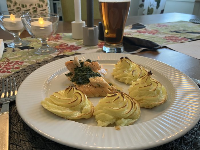 Middag med lax och duchessepotatis på tallrik, ölglas och tända ljus i bakgrunden. Estetiskt och inbjudande.
