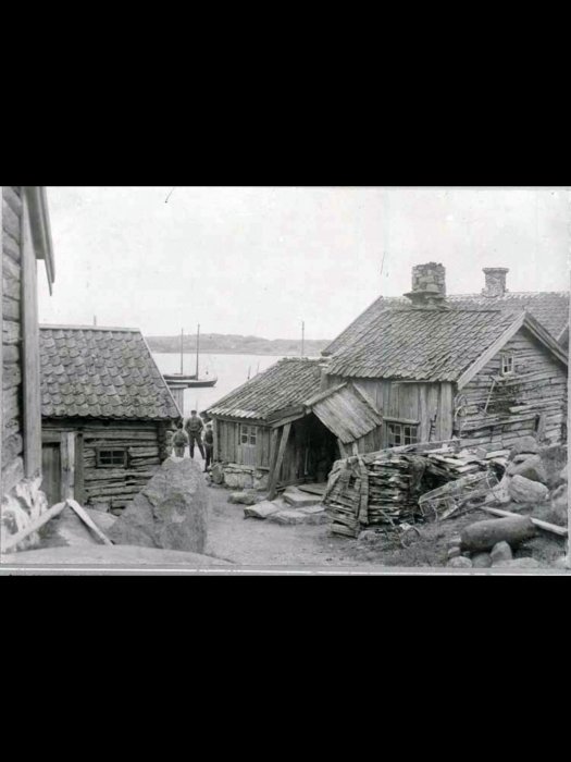 Svartvit bild, gammal fiskeby, trähus, stenig väg, båtar i bakgrunden, historisk atmosfär, några människor syns.