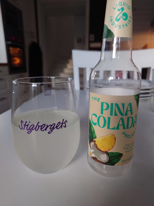 Ett glas med dryck och en Pina Colada-ciderflaska på ett bord, inomhusmiljö med ugn i bakgrunden.