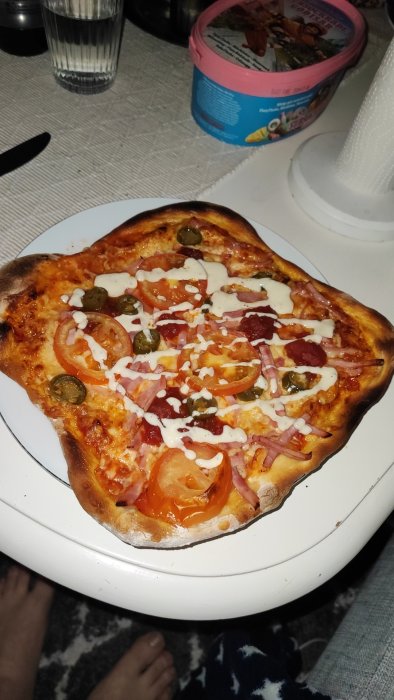 Hembakt pizza med skinka, tomater, jalapeños, vit sås, på vit tallrik, intill barfota fötter och glassförpackning.