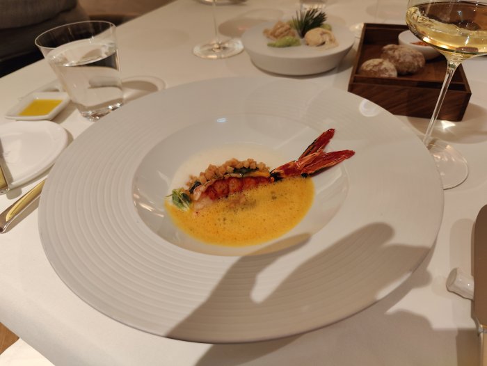 Gourmeträtt med räka, sås, örter på tallrik i fin restaurang, vin, bröd, vatten i bakgrunden.