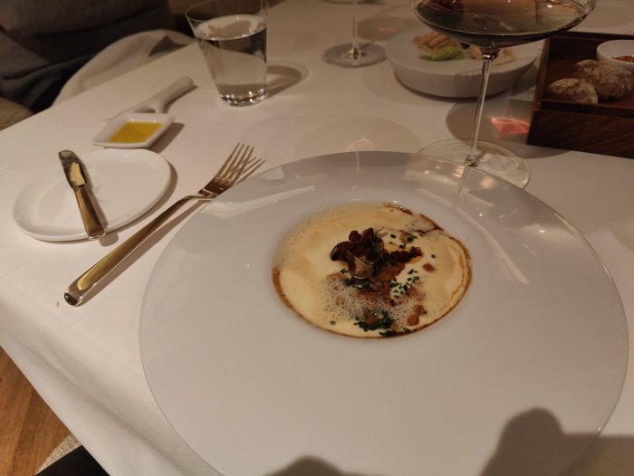 Fint dukat bord med maträtt, sås, champinjoner, skum, vit tallrik, restaurangmiljö, vinglas, bröd, vatten.
