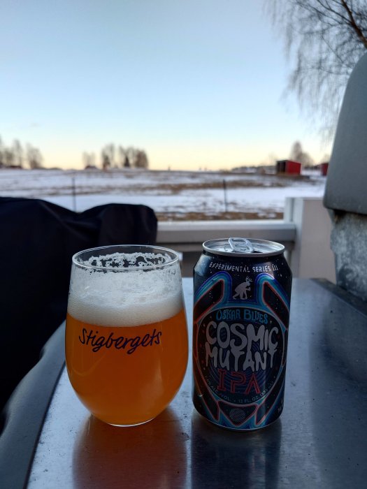 Ett glas öl och burk med etikett "COSMIC MUY THAI IPA" utomhus, vinterlandskap i bakgrunden, vid skymningen.