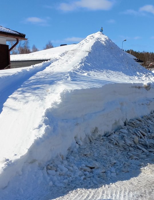 Stor snöhög mot klarblå himmel, byggnader i bakgrunden, soligt väder, isskulpturstruktur i förgrunden.