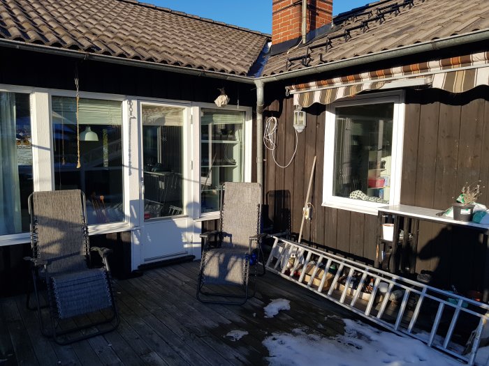 Trädäck med stolar, inglasad veranda, träsiding, snörester, soligt, skoträd, avslappnad utomhusmiljö.